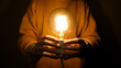 © tanitost - Hands holding glowing light bulb in front of torso on dark background. A pair of hands are gently holding a brightly glowing light bulb, symbolizing a breakthrough solution, a flash of inspiration