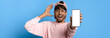 © Prostock-studio - A young man wearing a pink sweater and cap smiles broadly while holding up a smartphone. He appears excited and surprised, with a bright blue background enhancing the joyful scene.
