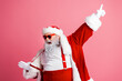 © deagreez - Santa in a red suit with white fur trim points up while wearing red suspenders and shades in a pink studio backdrop for a festive photo shoot