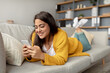 © Prostock-studio - A charming Arab lady lies comfortably on a sofa, using her smartphone to check social media and chat online. She looks happy and engaged while enjoying her time at home.