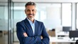 © Ger - A smiling, middle-aged man with gray hair and a beard poses with crossed arms in a modern office. He wears a blue suit