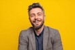 © Vitalii Shkurko - A man with a beard and gray blazer smiles cheerfully while sitting against a vibrant yellow backdrop. His expression shows warmth and friendliness, creating a joyful atmosphere
