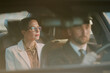 © AnnaStills - Caucasian middle aged woman sitting in back seat of car being driven by Caucasian middle aged man wearing suit and cap, both looking forward, business travel concept
