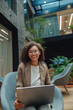 © Yaroslav Astakhov - diverse female freelancer smiling with laptop in cozy atrium armchair, relaxed posture, remote work setup,