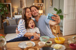 © Stockphotodirectors - A joyful family gathers around a table filled with food. They take a playful selfie, smiling happily as they bond together at home during meal time.