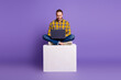© deagreez - Young handsome guy sits cross legged on a white block with a laptop in a purple studio background for casual fashion lifestyle advertising and shopping concepts