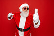 © InsideCreativeHouse - Crazy santa claus with grey beard wireless modern headset headphone holding smart phone looking at mobile screen listen music dance wear sunglass cap isolated bright shine red color background