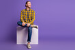 © deagreez - Young handsome man sits on white cube wearing yellow plaid shirt jeans sneakers purple background lifestyle fashion advertising