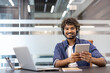 © Tetiana - Portrait of a young Indian man in a headset sitting in an office at a table with a laptop, holding a tablet in his hand and looking at the camera with a smile