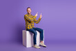 © deagreez - Young handsome man in casual checkered shirt sits on a white block against purple background smiling and pointing up for lifestyle advertising