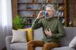 © Liubomir - Mature man with white beard clutching his chest on a living room sofa during an asthma attack, using a blue inhaler for urgent breathing relief and medical treatment
