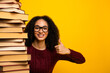© deagreez - Young woman with glasses smiles and stands beside a tall stack of book giving a thumbs up against a bright yellow background