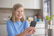 © Liubomir - Mature woman happily engaging with her smartphone, browsing the internet or connecting with family and friends remotely, while relaxing in her bright kitchen at home