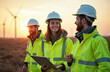 © miss irine - Three engineers in hi vis jackets and helmets work as a team at wind farm with turbines in background. Woman holds tablet while man talks, sun sets.