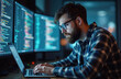 © miss irine - Young bearded man with glasses codes on laptop. Monitors display lines of computer programming code. Focused programmer works late night in dark office.