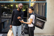 © DC Studio - African American client paying with his smart watch at the pos, accepting a contactless payment from a customer. Electronic transaction finalizing the takeout delivery exchange.