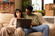 © DC Studio - Couple sits on couch with laptop in living room surrounded by moving boxes, browsing online together. Caucasian woman and black man discuss plans for new home while searching for ideas.