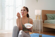 © eggeeggjiew - Asian woman in yoga outfit sitting on mat, happily eating fruit yogurt after workout in bright home morning light.