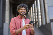 © Tetiana - Portrait of a young Indian man in a white shirt standing outside, holding a mobile phone and smiling at the camera