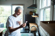 © Geber86 - Senior man smiling at smartphone while drinking water in home kitchen