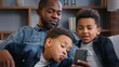© Yuliia - African American happy family with mobile phone smiling taking picture selfie grimacing father two kids sons boys children looking in smartphone trying funny photo mask recording video call at home