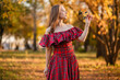© Вероника Преображенс - A young woman stands in an autumn forest. She is wearing a long, red, plaid dress with off-the-shoulder details and a frill, a romantic, vintage style. She holds a decorative metal flower in her hands