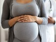 © Thanagorn - A pregnant woman in a gray outfit holds her belly, showcasing her pregnancy, while a doctor stands in the background, indicating a medical setting.
