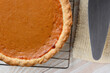 © Steve Cukrov - Closeup of a fresh baked pumpkin pie on a cooling rack next to a serving spatula and towel. Horizontal format on a white rustic wooden kitchen table.