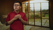 © Krakenimages.com - Man young hispanic clasping hands in a building room by large window, wearing red polo and glasses; introspection solitude.