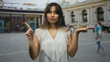 © Krakenimages.com - Young latina woman points index finger with one hand and holds other palm up in front of an ornate building on a busy street; doubt choice decision.