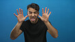 © Krakenimages.com - Young hispanic man making playful faces against isolated blue background wearing black shirt with expressive gestures and humorous expressions showcasing personality.