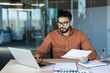 © Liubomir - Businessman sitting at a desk in a modern office, working on a laptop while holding and reviewing paper documents, demonstrating dedication and productivity