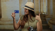 © Krakenimages.com - Woman smiling with credit card at historic university, wearing hat and camera, gesturing approval in sunny courtyard, celebrating travel and exploration in cultural setting.