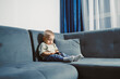 © DSMT - Curious toddler explores smartphone while sitting comfortably on a blue couch in a sunlit living room