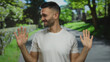 © Krakenimages.com - Young man gesturing happily in a lush green park setting under bright sunlight, conveying a carefree and joyful vibe with trees and path in the background.