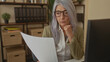 © Krakenimages.com - Senior woman with grey hair in an office setting, thoughtfully analyzing documents at her desk, surrounded by shelves and workplace essentials.