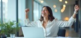 Woman celebrates success at laptop. She smiles widely with arms raised in office. Financial freedom or crypto trading gains achieved. Happiness and winning at work.