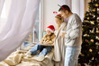 © Studio Romantic - Christmas family by window, child in Santa hats. Parents hold mugs while reading a book with child girl in a warm room by festive tree at home. Winter holiday mood and New year celebration.