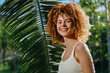 © SHOTPRIME STUDIO - smiling woman with curly hair wearing white tank top stands outdoors near tropical palm leaves in warm sunlight, natural beauty and happiness captured.