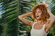 © SHOTPRIME STUDIO - Smiling woman with curly hair enjoys sunny tropical outdoors surrounded by large green palm leaves, wearing white tank top and expressing relaxed happiness.