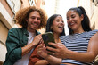 © Ladanifer - Three happy multiracial young friends are having fun together outdoors, enjoying good time and using mobile phone