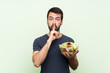 © luismolinero - Young handsome man with salad over isolated green wall showing a sign of silence gesture putting finger in mouth