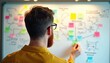 © miss irine - Bearded man works on whiteboard planning user experience. He organizes colorful sticky notes to map out a tech project, digital design, and business strategy. He develops new product processes.