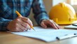 © miss irine - Person in a blue plaid shirt signs construction documents. Yellow pen and hardhat on table. A home renovation contract is underway. Business deal or agreement about the house.