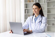 © Anastasiya - Smiling female doctor in white coat working on laptop in bright clinic office, professional healthcare specialist using technology for medical consultation