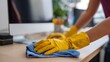 © imagemir - A woman in yellow gloves cleaning an office desk with a blue cloth, focused and detail-oriented in her task.
