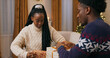 © ABCreative - A brother and sister of multi-racial background sit on a sofa in warm sweaters, packing presents for friends. The young woman shows her brother how to tie a ribbon. They communicate and smile on the