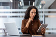 © Liubomir - Professional businesswoman in smart casual attire reviewing data on a digital tablet at her desk with a laptop, focused and productive in a modern hybrid office workspace