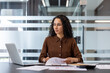 © Liubomir - Young businesswoman with curly hair sitting at her office desk, looking focused while working with a laptop and analyzing paperwork and financial reports