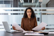 © Liubomir - Stressed businesswoman sitting at a modern office desk, looking directly at the viewer with a concerned expression while holding papers, signifying workload, financial problems, and confusion